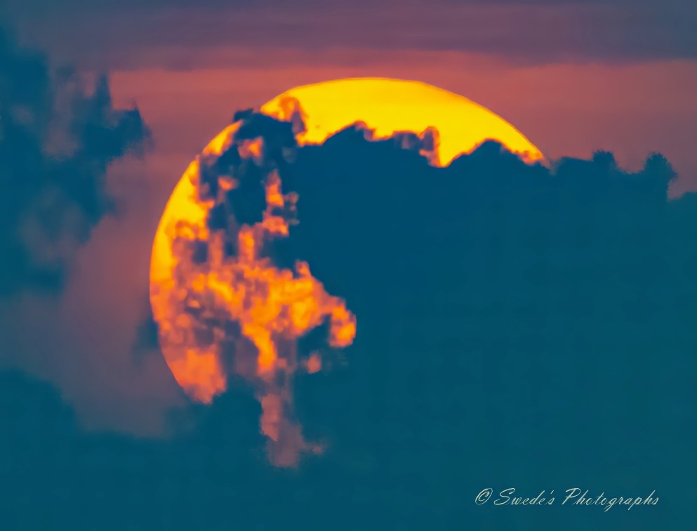 "The image shows a large, bright, orange-yellow sun partially obscured by dark clouds. The sun is positioned in the center of the image, with the clouds creating an interesting silhouette against the vibrant background. The sky around the sun has a gradient of colors, transitioning from deep blue to lighter shades of orange and pink. The contrast between the dark clouds and the bright sun creates a dramatic and visually striking scene. The image is signed with the text "© Swede's Photographs" in the bottom right corner." - Copilot