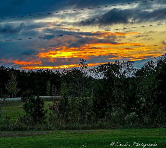 "The image shows a scenic landscape during sunset. The sky is filled with dramatic clouds, with vibrant hues of orange and yellow from the setting sun peeking through. The foreground features a grassy field with some bushes and trees, while the background includes a line of trees and a road. The contrast between the dark clouds and the bright sunset creates a visually striking and serene scene. The image is signed with "© Swede's Photographs" in the bottom right corner.

It’s a beautiful moment captured, blending the tranquility of nature with the dramatic colors of a setting sun." - Copilot