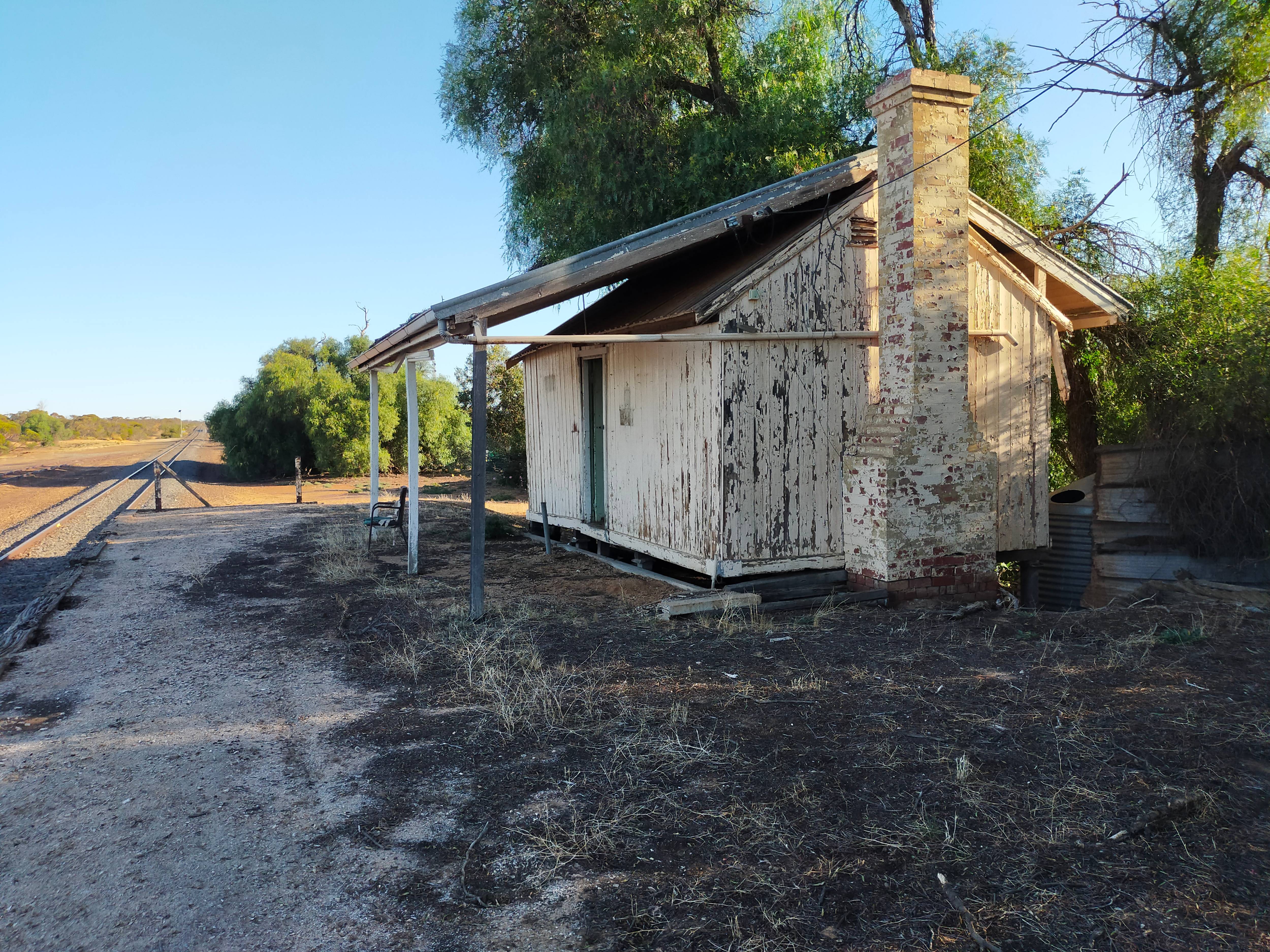 A dilapidated, yellowish-white, wooden building with flaking paint on a half overgrown gravel platform at Hattah Victoria