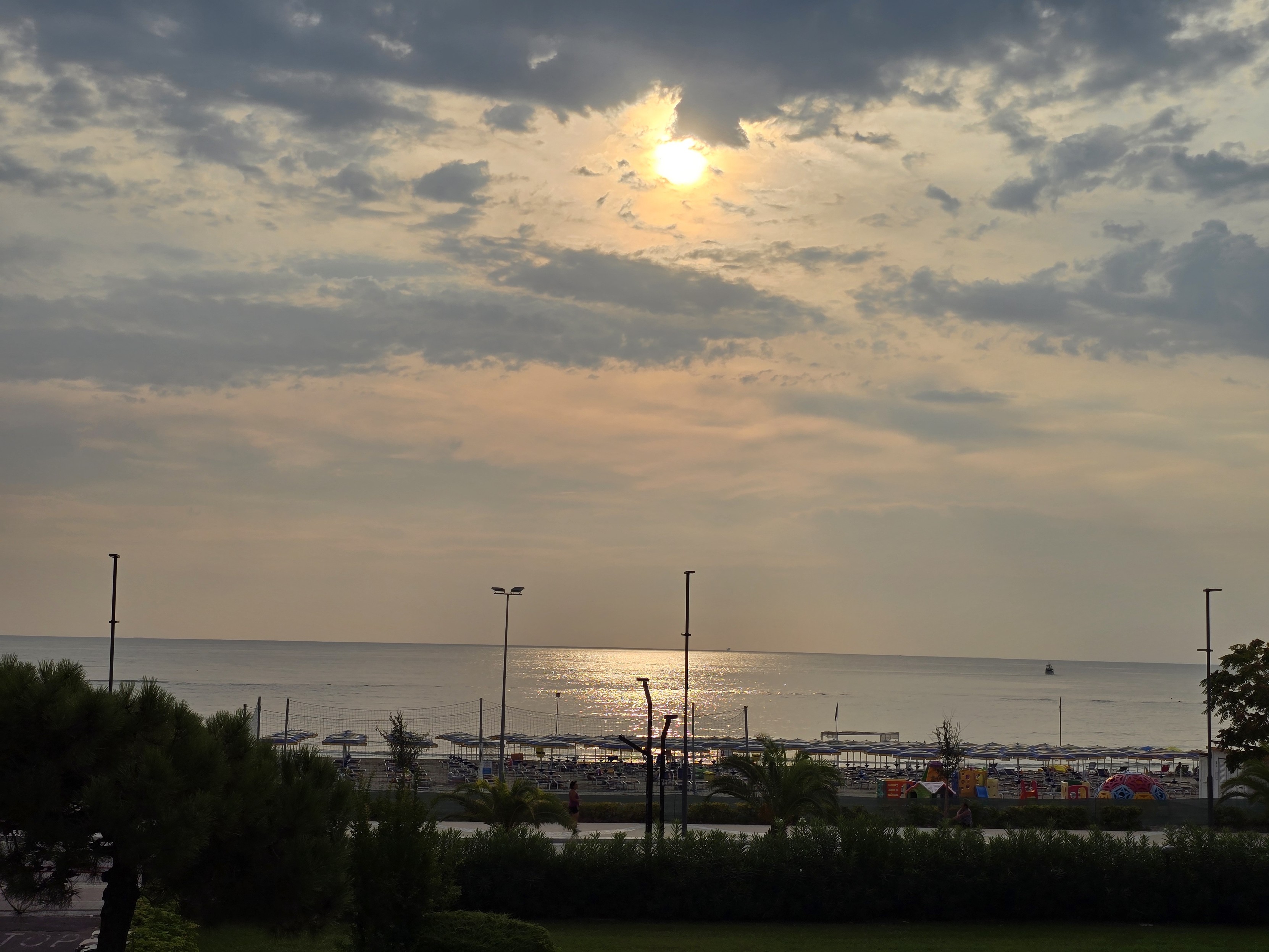 A serene sunrise casts a warm golden glow across the sky, with soft sunlight filtering through gentle clouds. The calm sea below reflects the shimmering light of the rising sun, creating a peaceful, mirrored effect. In the foreground, a beach dotted with empty sunbeds and umbrellas rests quietly, with only a few figures strolling by the shore. The silhouettes of trees and lampposts frame the scene, enhancing the tranquil atmosphere of this early morning coastal view. The muted hues of the sky and water suggest the start of a calm, beautiful day.