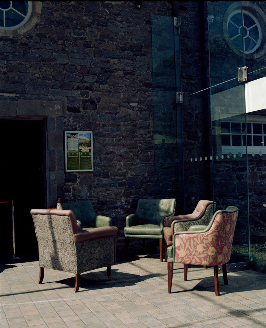 a photo of a group of chairs upholstered in leather and patterned cloth, arranged in an informal circle as if a group has been sat talking in them. The chairs are empty now, and late-date sunlight, before golden hour, pours onto them. The chairs are in a glass walled area between two old buildings, with the original stone exterior next to the chairs, now included inside the glass seating area. The walls have round, single-pane glass windows, and a small poster of announcements