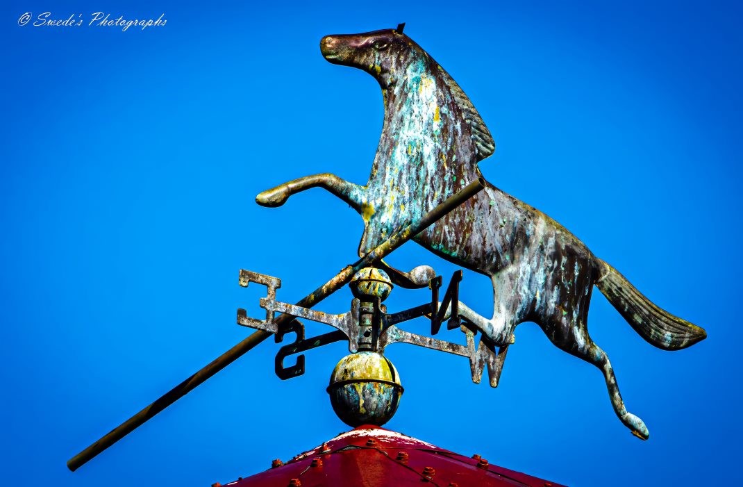 "The image shows a weathervane featuring a horse in mid-gallop, mounted on top of a red structure, likely the peak of a roof. The weathervane is set against a clear blue sky. The horse figure appears weathered, with peeling paint and signs of oxidation on the metal beneath. This combination of functionality and decorative art highlights the effects of time and exposure to the elements on such objects." - Copilot