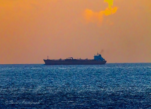 "The image shows a large ship on the horizon at sea during either sunrise or sunset. The sky and water are painted with beautiful orange and blue hues, creating a serene and picturesque scene. The ship is silhouetted against the vibrant colors, with visible details like its structure and what appear to be cranes or masts. The overall mood is peaceful and solitary, capturing the essence of maritime travel." - Copilot