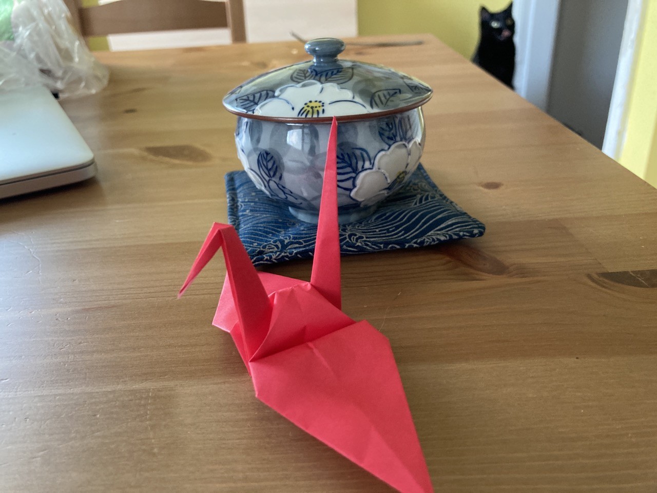 A bright red crane origami. a black cat has her goofy pink tongue sticking out in the background. 