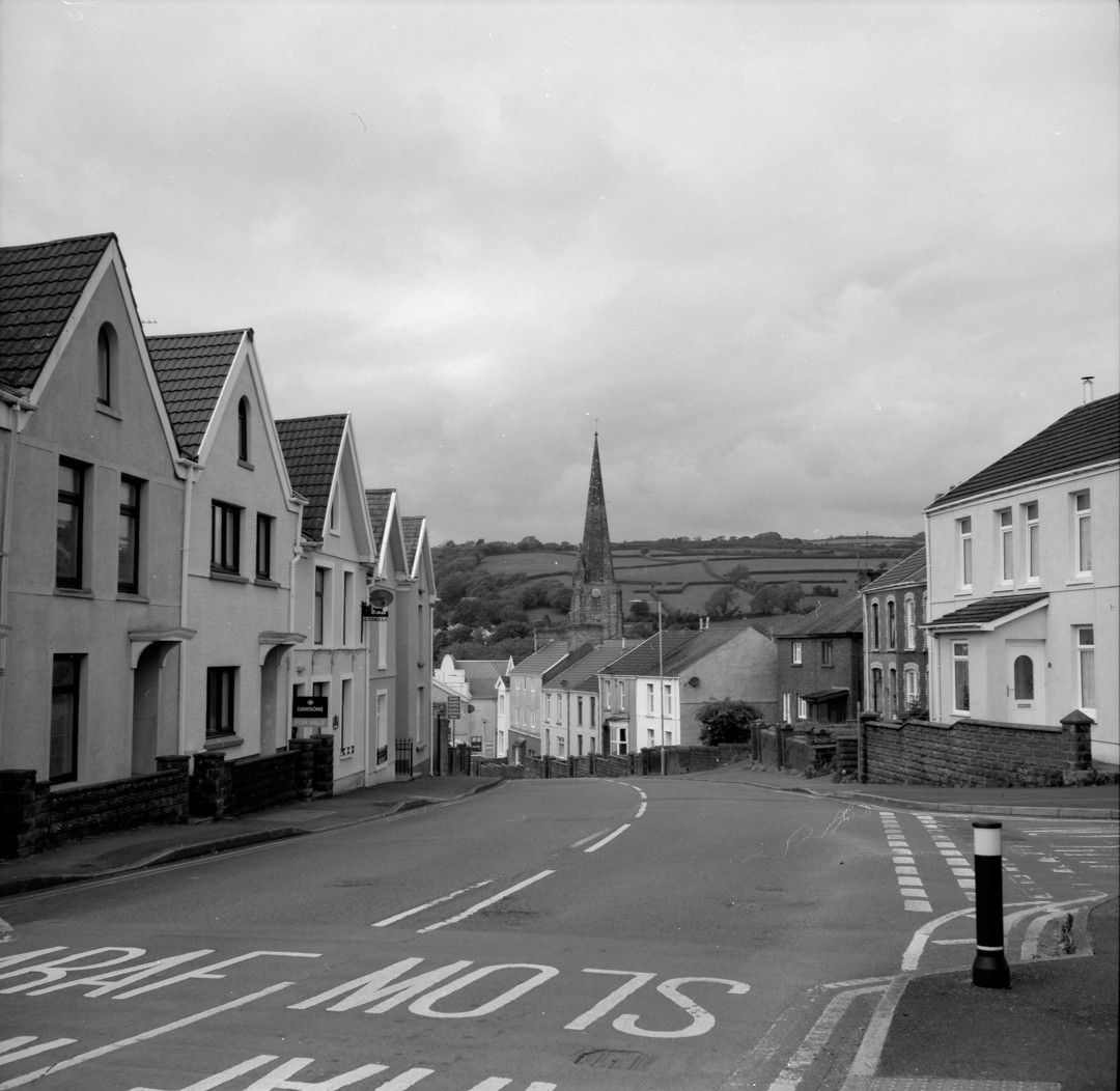 a black and white photo of an empty, two lane road leading between terraced houses. the road curves across the frame, from the left corner to the centre. in the distance a spire is visible with a clock on the side, and hills in the far distance