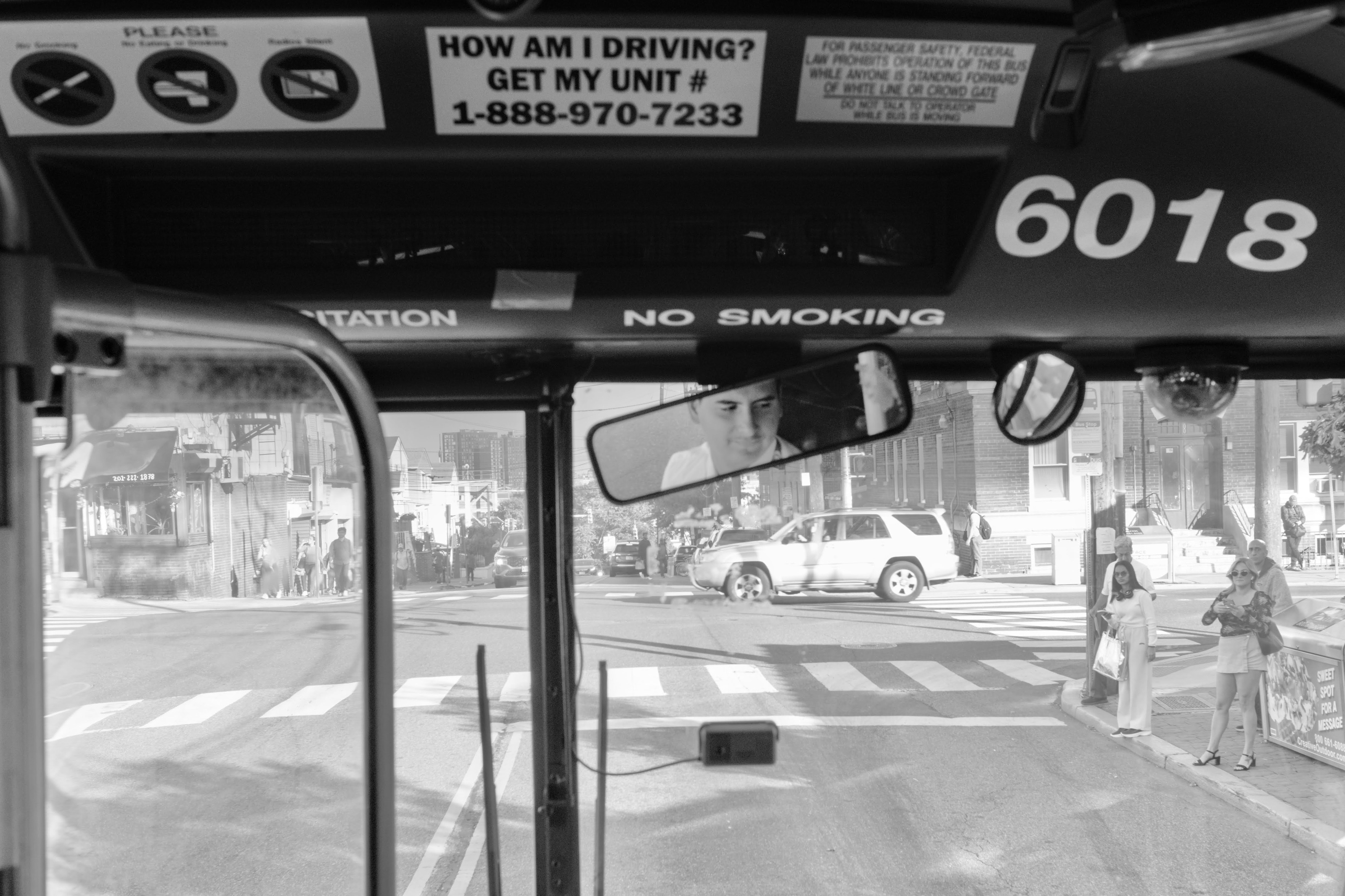 A black and white photo from inside the front of a 119 bus in Jersey city, looking out into a busy intersection. The driver is seen in the rear view window reflection. 