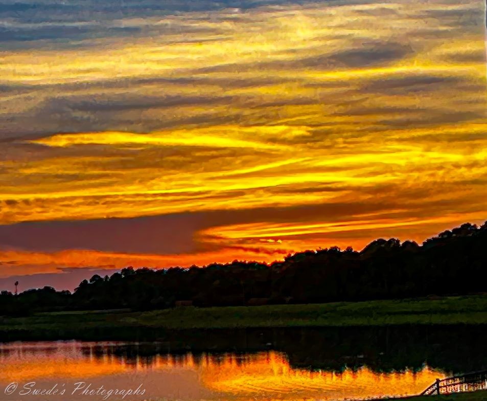 "The image shows a vibrant sunset over a body of water. The sky is filled with dramatic, colorful clouds in shades of yellow, orange, and red, creating a striking contrast with the darker silhouette of the trees and landscape below. The water reflects the vivid colors of the sky, enhancing the overall beauty of the scene. The image is interesting due to the intense and varied colors of the sunset, the reflection on the water, and the peaceful, natural setting it depicts." = Copilot