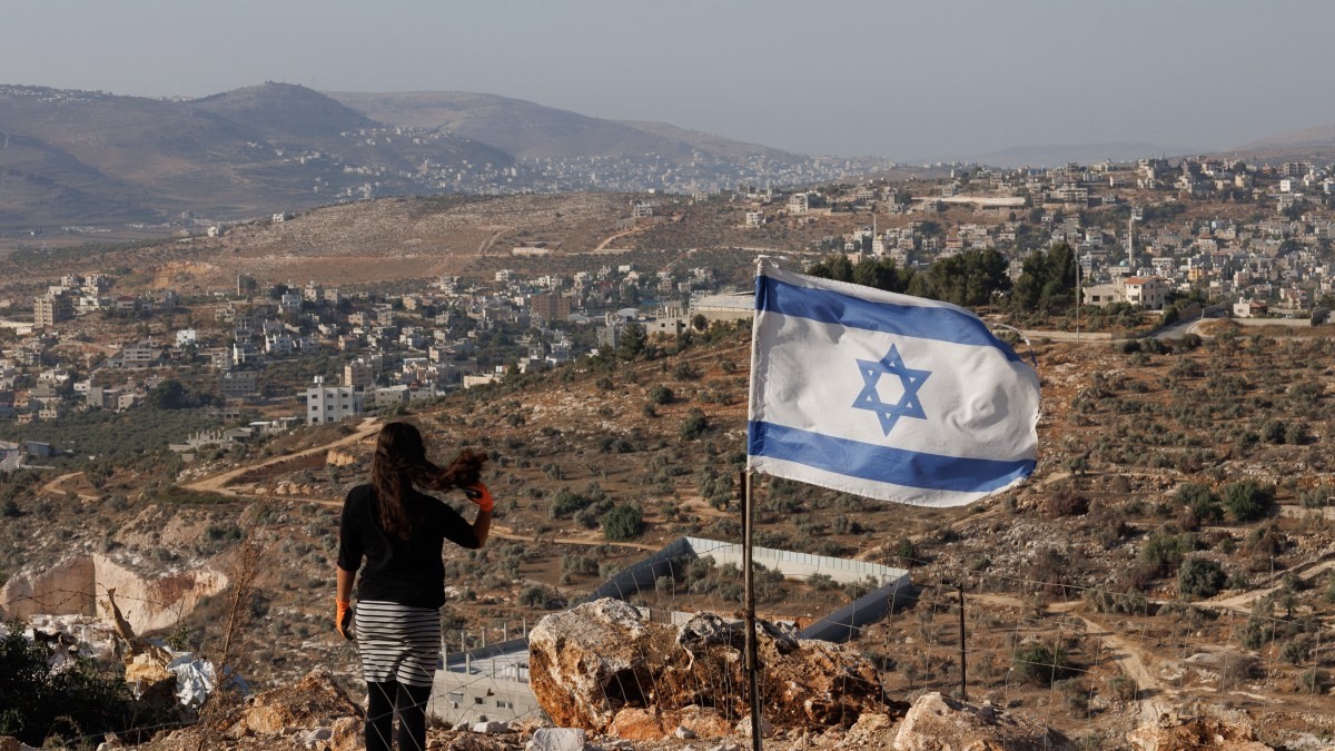 Photo: a woman stands by an Israeli flag looking down on stolen land

Caption An Israeli settler above the Palestinian West Bank village of Beita in the illegal outpost Evyatar