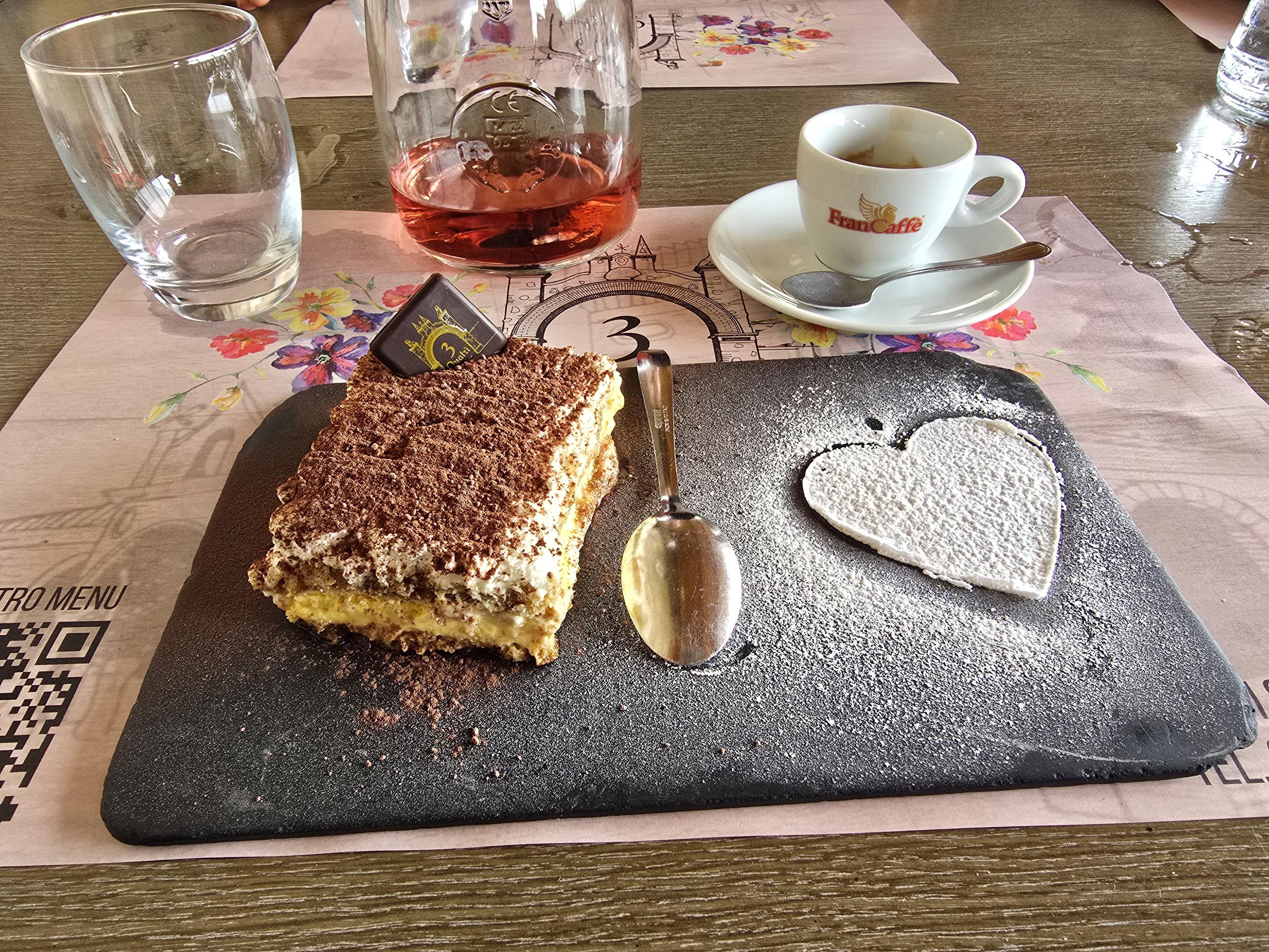 The image shows a table set with a dessert and drinks. A slice of tiramisu is served on a rectangular dark slate plate, dusted with cocoa powder. Next to the tiramisu, there's a powdered sugar heart-shaped decoration. A small square of dark chocolate is placed on the tiramisu. A spoon rests on the plate. To the side, there’s a glass of cerasuolo wine, a glass of water, and an espresso cup with a spoon placed on the saucer. The table mat underneath has a floral design, and there is a QR code visible in the bottom left corner of the mat.